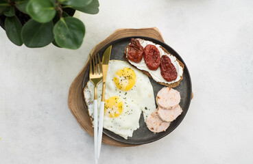 Balanced breakfast of two fried eggs, sandwich of cream cheese and dried tomatoes, slices of poultry meat on gray background.