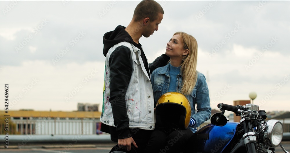 Wall mural A young stylish couple of bikers meet and hug near a motorcycle standing in a public city parking lot