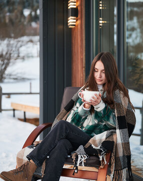 Young woman resting on terrace of modern barn house in the mountains. Happy female tourist sitting in chair, holding cup of tea, enjoying in new cottage in winter.
