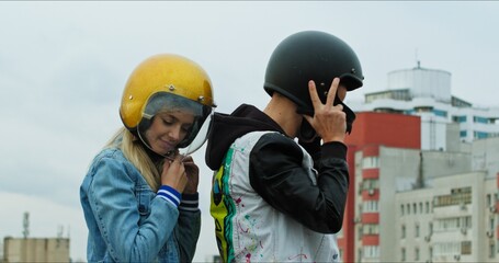 A young stylish couple of bikers sit on a bike standing in a public city parking. The guy and the girl put on motorcycle helmets. Love and safety