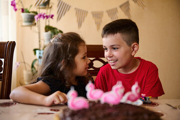 Charming Caucasian kids, a teenage boy and elementary age girl, brother and his younger sister having fun together, sitting at a table with blurred chocolate birthday cake with pink flamingo candles