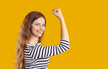 Happy confident beautiful young woman standing isolated on yellow background, looking at camera, flexing her strong arm and smiling. Side profile view. Girl power, self confidence, empowerment concept
