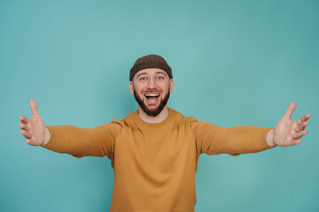 Lets hug! Handsome beardy young caucasian man in yellow sweater smiling wide spreading hands in welcome gesture. Cheerful Italian male welcoming person isolated over turquoise studio backdrop. Mockup.