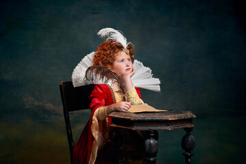 Portrait of little red-headed girl in costume of royal person writing letter with thoughtful look isolated over dark green background