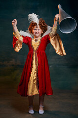 Portrait of little red-headed girl, child in costume of royal person isolated over dark green background. Winning look