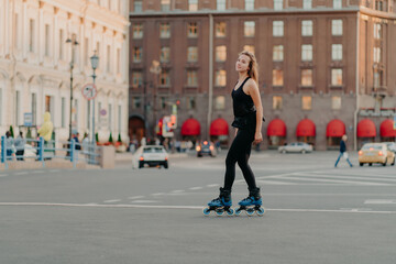 Full length shot of slim dark haired woman rollerblades on asphalt road dresed in black sportsclothes enjoys outdoor activities exercises for healthy and strong body. Sport and hobby concept