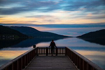 Obraz premium dark silhouette of a girl standing on a pier over the sea with a reflection of a unique colourful sunset, sunset on lofoten islands in norway