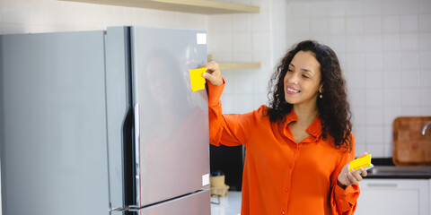 Young and organized female with curly hair in casual clothing making to-do and checklist on sticky notes as reminder and placing them on refrigerator at home