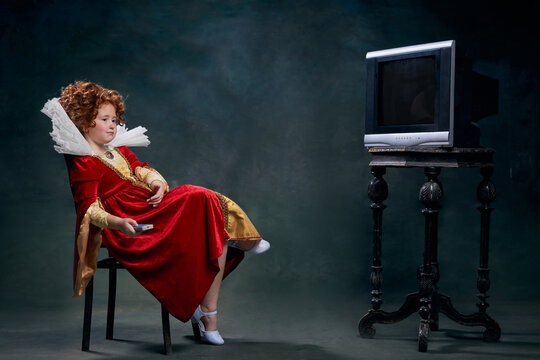 Portrait Of Little Red-headed Girl, Child, In Image Of Royal Person Sitting In Front Of TV Isolated Over Dark Green Background. Movie Time