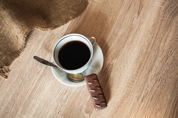 A cup of fragrant black coffee on a saucer with a spoon and a chocolate bar lie on a wooden brown table covered with linen.
