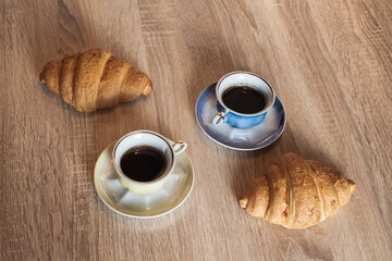 A couple of cups of hot fragrant black coffee in a saucer with croissants on a light brown wooden table.