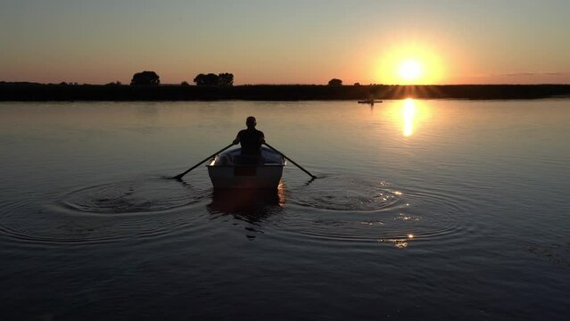 An elderly man with a poodle dog swims away from the camera on a boat at dusk in the late evening against the backdrop of sunset