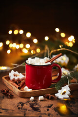 Red cup of hot cocoa with marshmallows on wooden table with christmas tree and glowing garland for...