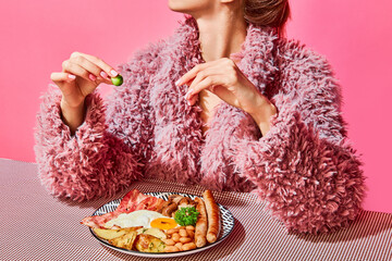Woman in furry pink coat eating English breakfast with eggs, bacon, sausage and vegetables