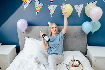 Happy emotional woman in pajama and party cap sitting on the bed with festive cake and champagne...