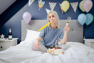 Sad, upset woman in pajama and party cap blowing in birthday pipe while sitting on the bed with...