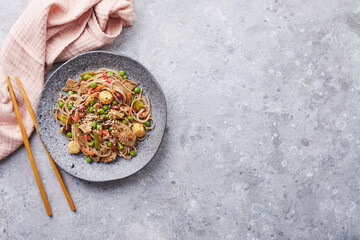 Wok with turkey meat, soba noodles, corn, green peas, green beans and carrots served on gray background with chopsticks. Asian food, concept of street food
