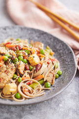 Wok with turkey meat, soba noodles, corn, green peas, green beans and carrots served on gray background with chopsticks. Asian food, concept of street food
