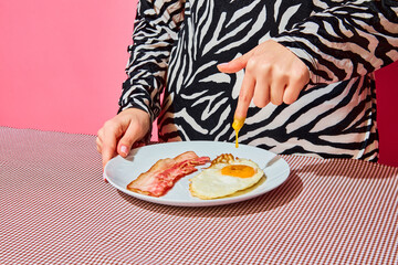 Woman putting finger inside egg yolk. Plate of English breakfast on pink tablecloth