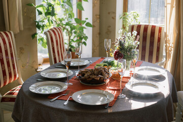 The table is set for a family dinner. Plates and glasses on a red tablecloth, meat and chicken with vegetables. Bouquet of flowers on the table