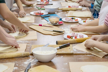 Long kitchen table with food ingredients and utensils, around which children stand and mold dough pieces for pizza. Creative process.
