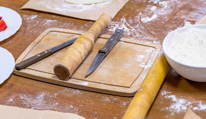 On wooden kitchen table sprinkled with flour are cutting board, knives, rolling pin, plate of flour.