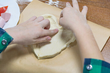On wooden kitchen table, child's cut hands form dough into small heart-shaped pizza.