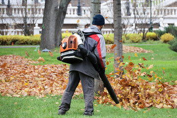 Man worker clean the city street with leaf blower. Falling leaves removal in autumn park