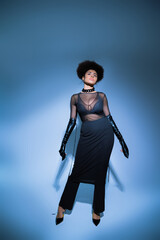 full length of curly african american woman in transparent shirt and black skirt looking up on blue.