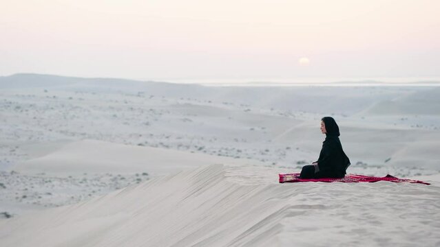 Young Arab muslim woman in traditional black hijab seated at carpet watching the sunset at the desert
