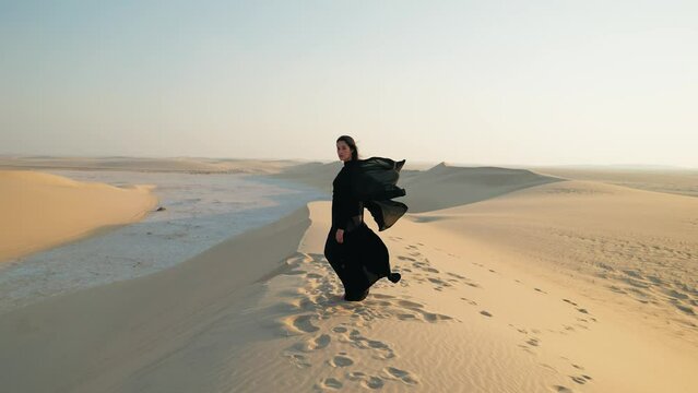 Young beautiful girl in traditional Abaya dress stand along the dunes in the desert of Qatar
