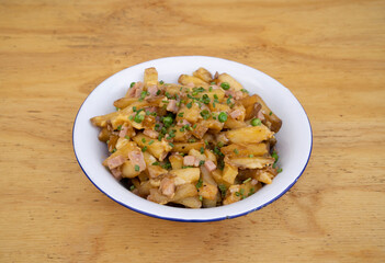 American food. Fried potatoes with egg, ham, peas and cheese, in a white bowl on the restaurant wooden table.