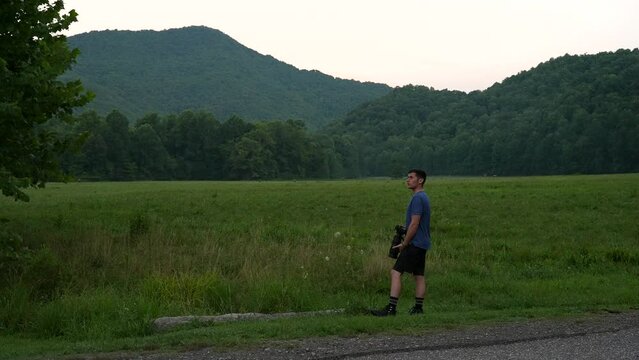 A wildlife photographer with a long telephoto lens looking for birds in the backdrop of a meadow and forested mountains