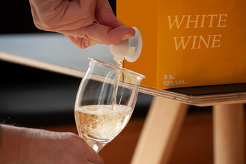 Hand pouring white wine into a glass from a BIB - cardboard bag in box with open tap standing on a table. Close up image. Fictitious brand. - 542995661