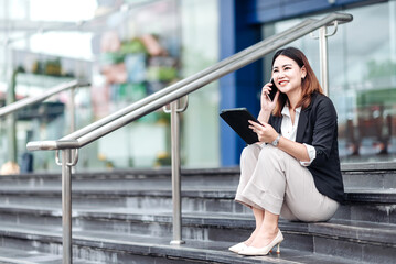 Young Asian businesswoman talking on phone and using tablet. Beautiful woman passenger has mobile call and discusses something with smile, holds coffee in hand