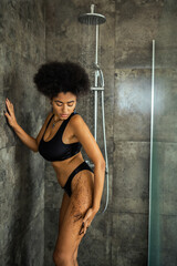 Curly african american woman applying coffee scrub under water in shower cabin.