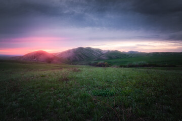 View of the hills and mountains covered by green grass under purple cloudy sky lightened by rising sun