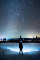 A man with a headlamp stands at night on a winter path under a bright starry sky and the Orion constellation