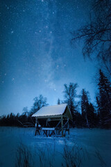 Mlky way in the night sky above snow-covered garden house in winter forest