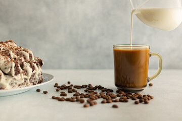 Coffee with cream in a glass mug with coffee beans, cream cake. cream is poured from a glass jug. Drink on a light gray background.