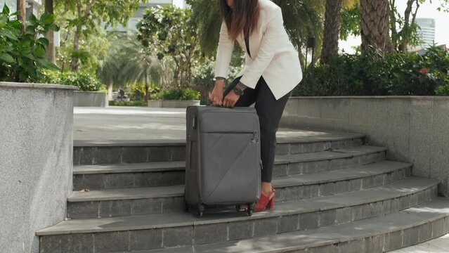 African American businesswoman having difficulties with pulling heavy suitcase while walking up stairs outdoors