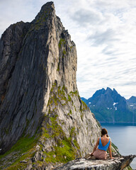 Fototapeta premium a beautiful girl sits on the rocks enjoying the view of the famous segla mountain in norway, senja island; hesten trail head, the famous norwegian fjords with mighty mountains above the sea
