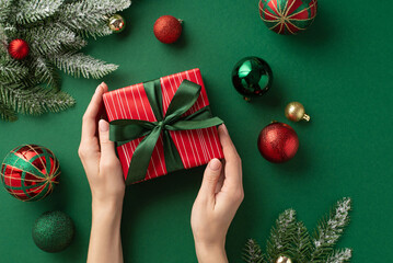Christmas Eve concept. First person top view photo of woman's hands holding red gift box with ribbon bow over green gold red baubles balls and pine branches in frost on isolated green background