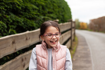 Fototapeta premium Little beautiful cute girl in glasses laughing near a wooden fence. Village child in a vest on a walk in the countryside