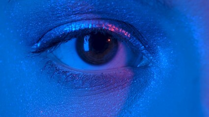 Womens eye with shiny make up with sparkles and arrow looking straight. Look of female eye with eyelashes and eyebrows illuminated by blue neon light. Macro portrait of female eye with brown iris.