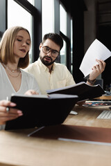 Businessman holding paper folder and talking to young intern with notebook in office.