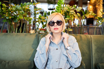 Stylish young blonde in sunglasses sitting at a table in a beautiful restaurant waiting for an order. The woman has a break in the working day