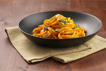 A deep black bowl with vegan pasta spaghetti carbonara with orange pumpkin sauce and fresh green herbs on kitchen towel on wooden background