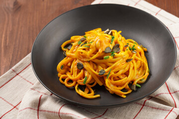 A deep black bowl with vegan pasta spaghetti carbonara with orange pumpkin sauce and fresh green herbs on kitchen towel on wooden background