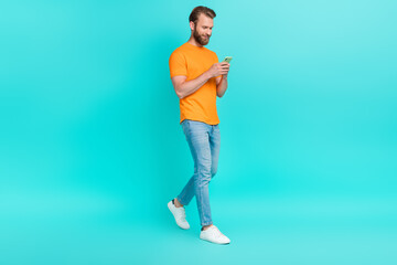 Fototapeta na wymiar Full size photo of handsome positive man dressed orange t-shirt walking read notification in smartphone isolated on teal color background